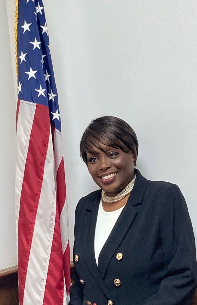 Myra Washington Dyson - Professional portrait in navy blazer with gold buttons and pearl necklace, smiling warmly with American flag backdrop